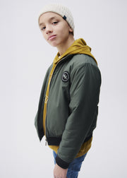 Boy wearing Mayoral Nukutavake forest green bomber jacket open over mustard yellow hoodie with cream beanie – on model side view