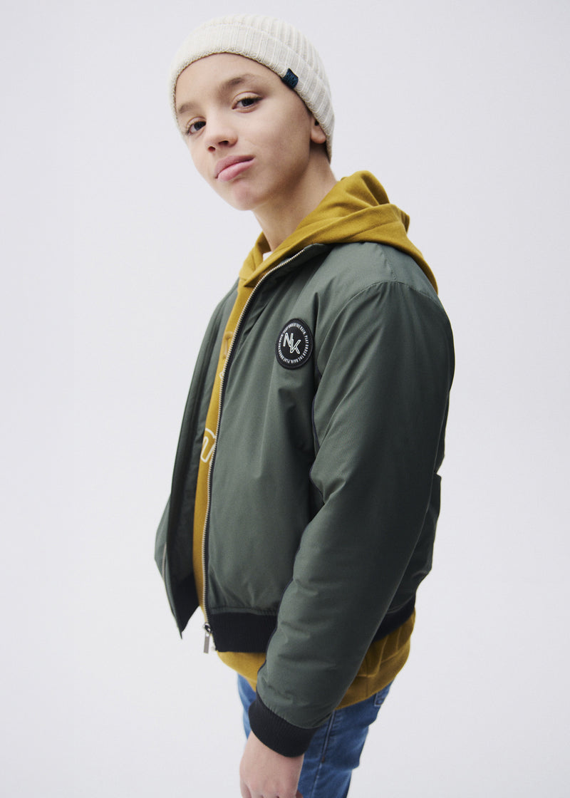 Boy wearing Mayoral Nukutavake forest green bomber jacket open over mustard yellow hoodie with cream beanie – on model side view